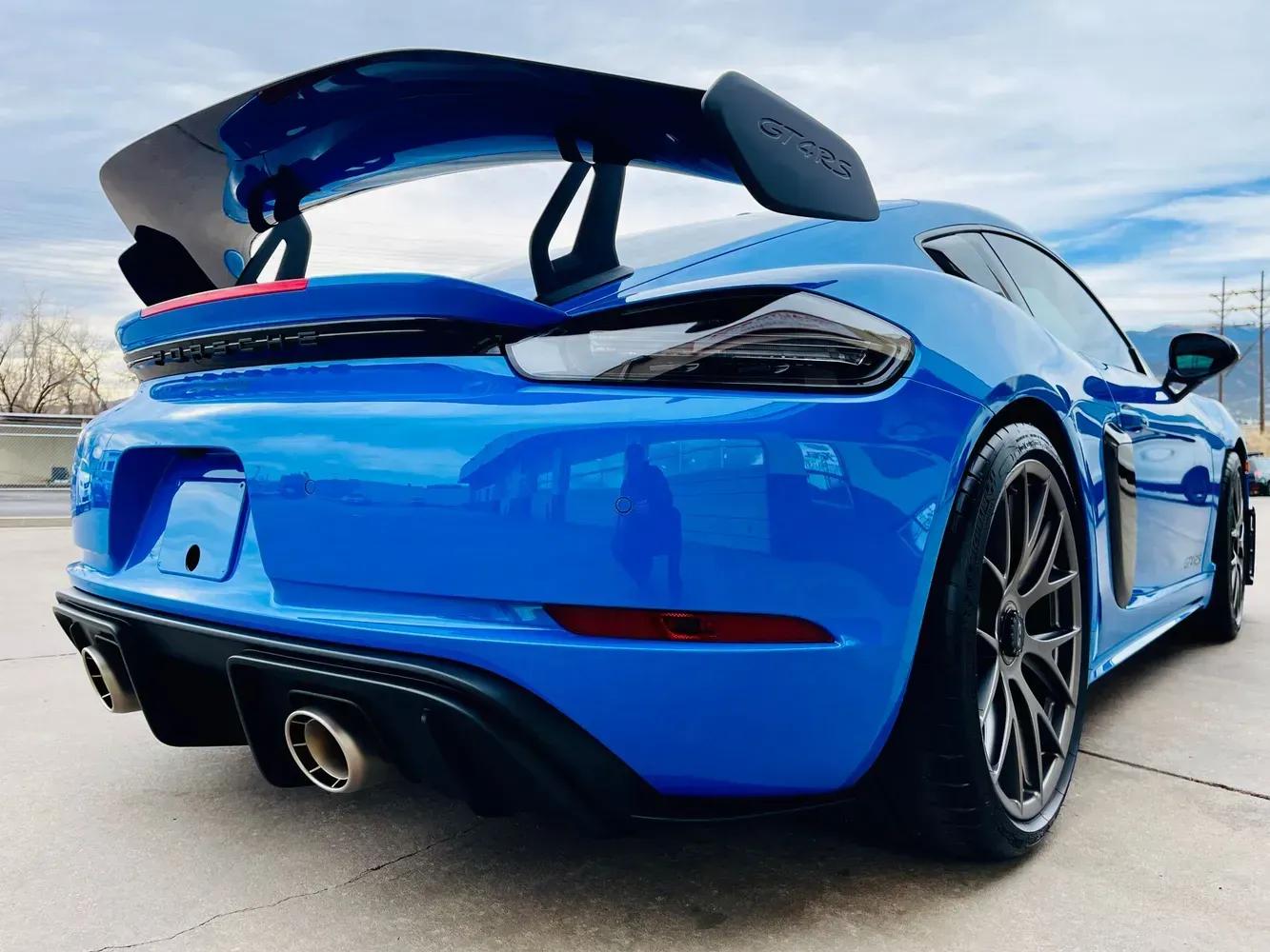 A bright blue Porsche 718 Cayman GT4 parked on a concrete lot, showcasing its rear spoiler, dual exhaust, and dark wheels.