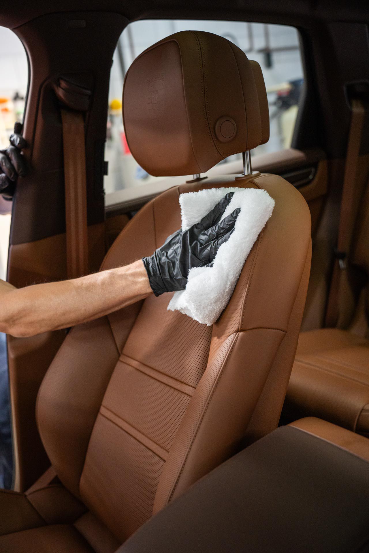 A person wearing black gloves cleans a tan leather car seat with a white cloth.