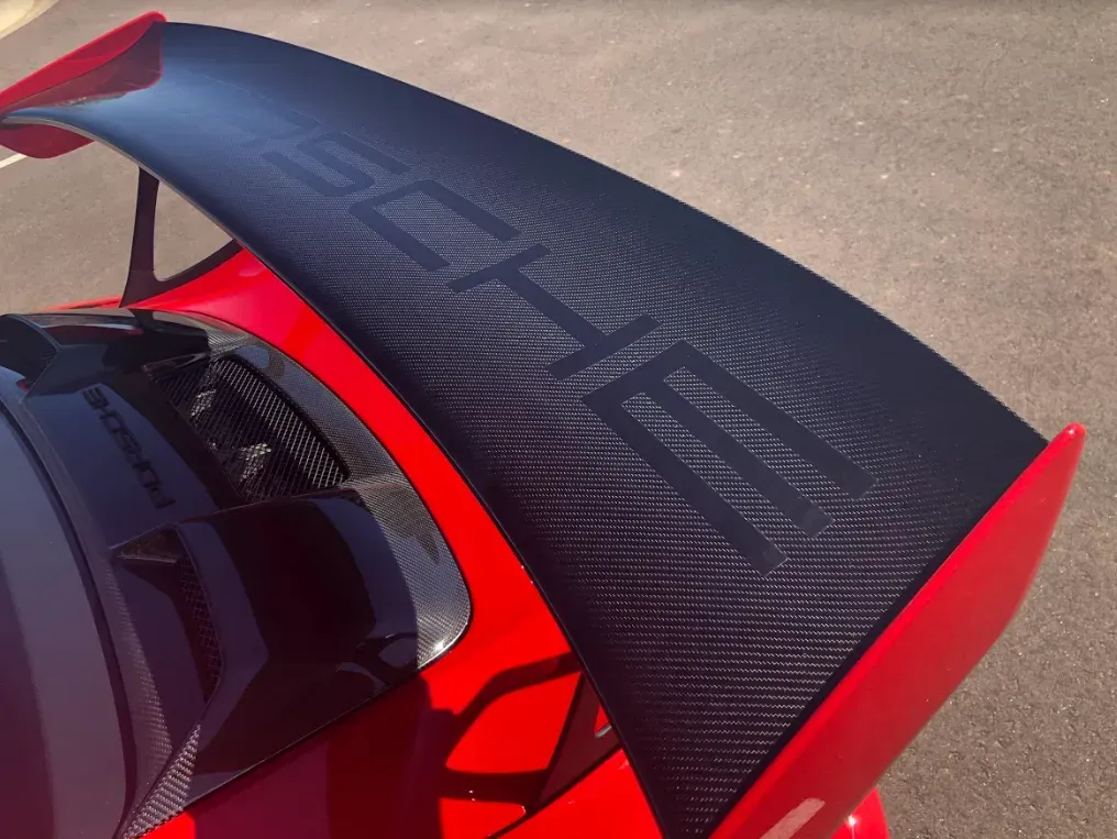 A close-up, high-angle shot of a bright red car featuring a large, black carbon fiber spoiler with PORSCHE lettering.
