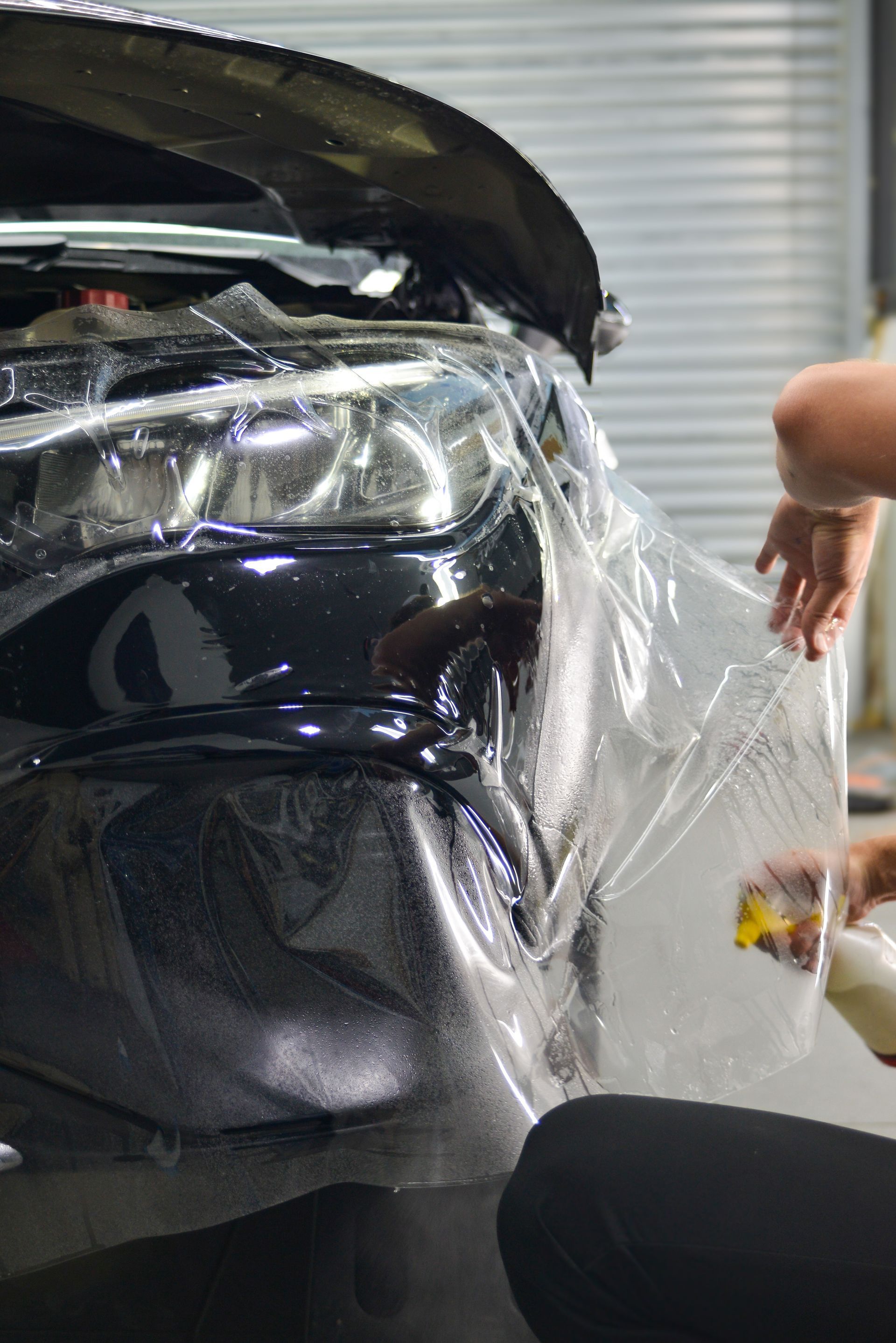 A technician applies a clear protective film to the front bumper and headlight of a black vehicle in a garage.