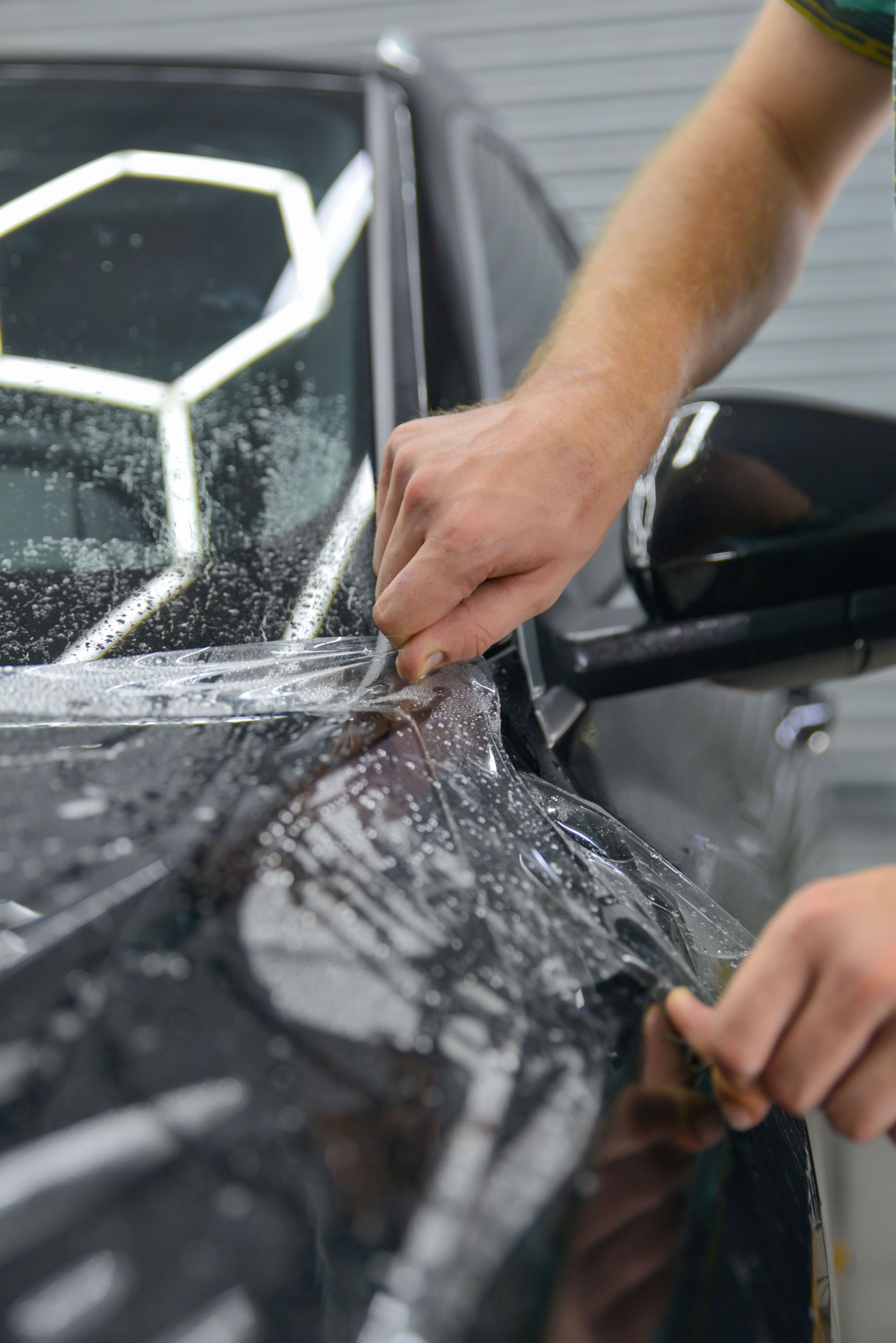 A technician uses a tool to carefully apply a protective film to the fender of a dark-colored car in a garage.