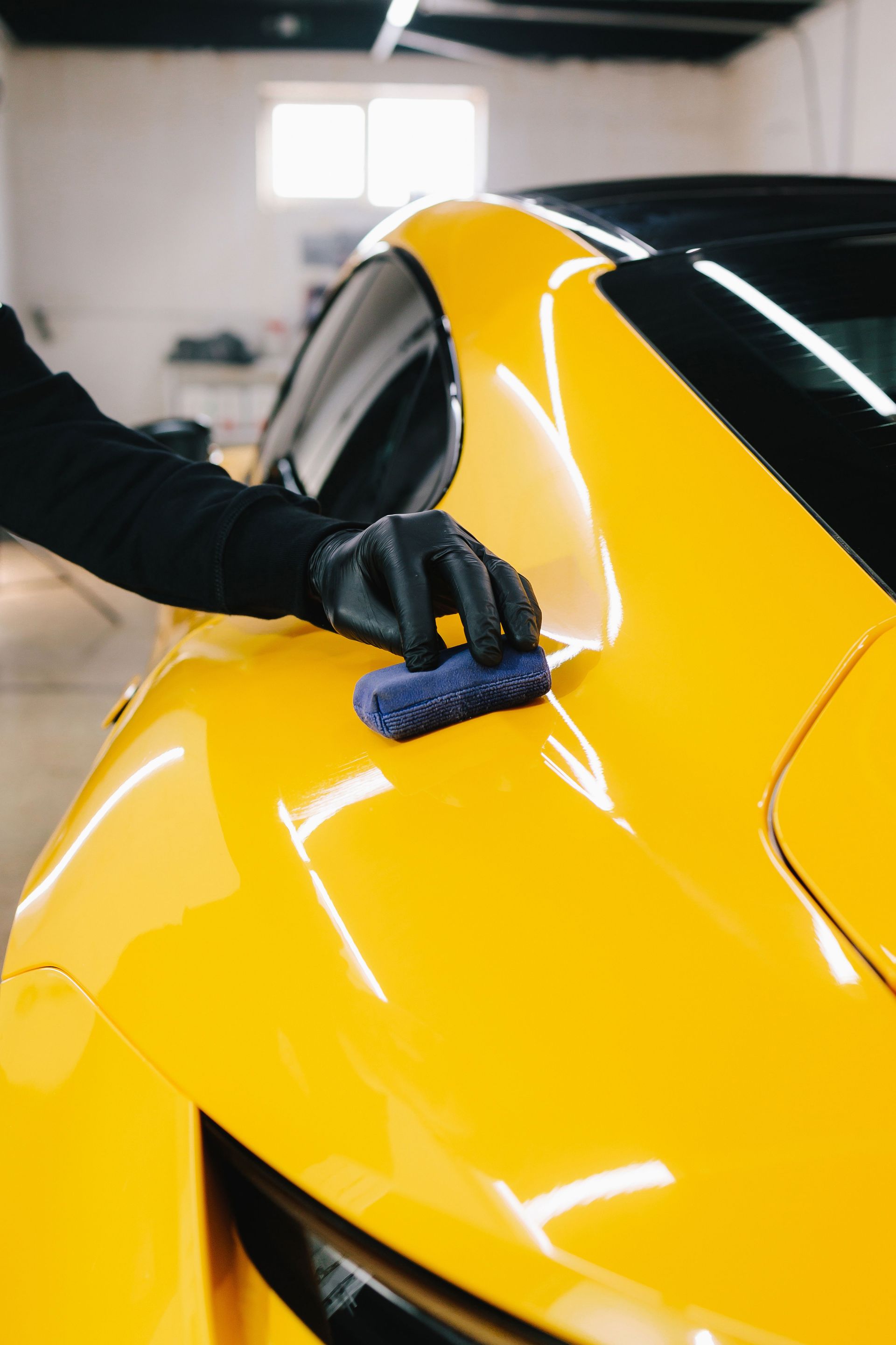 A person in a black glove uses a blue applicator sponge to apply a detailing product to the yellow body of a car.