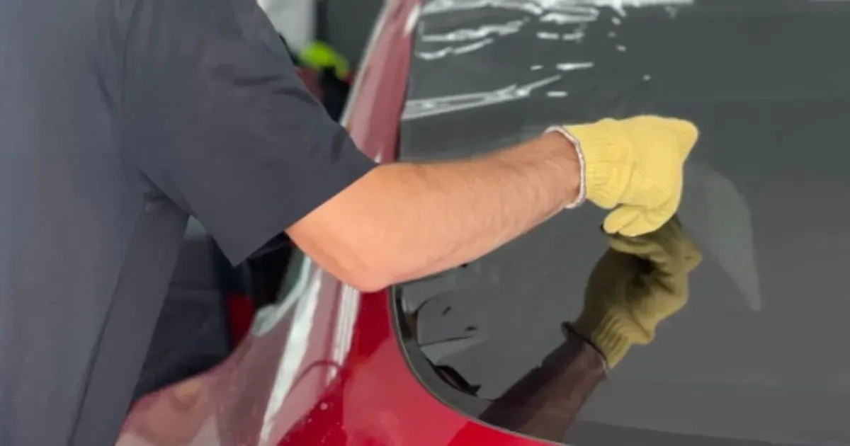 A worker wearing a yellow glove uses a squeegee to apply tinted film to a red car's rear window.