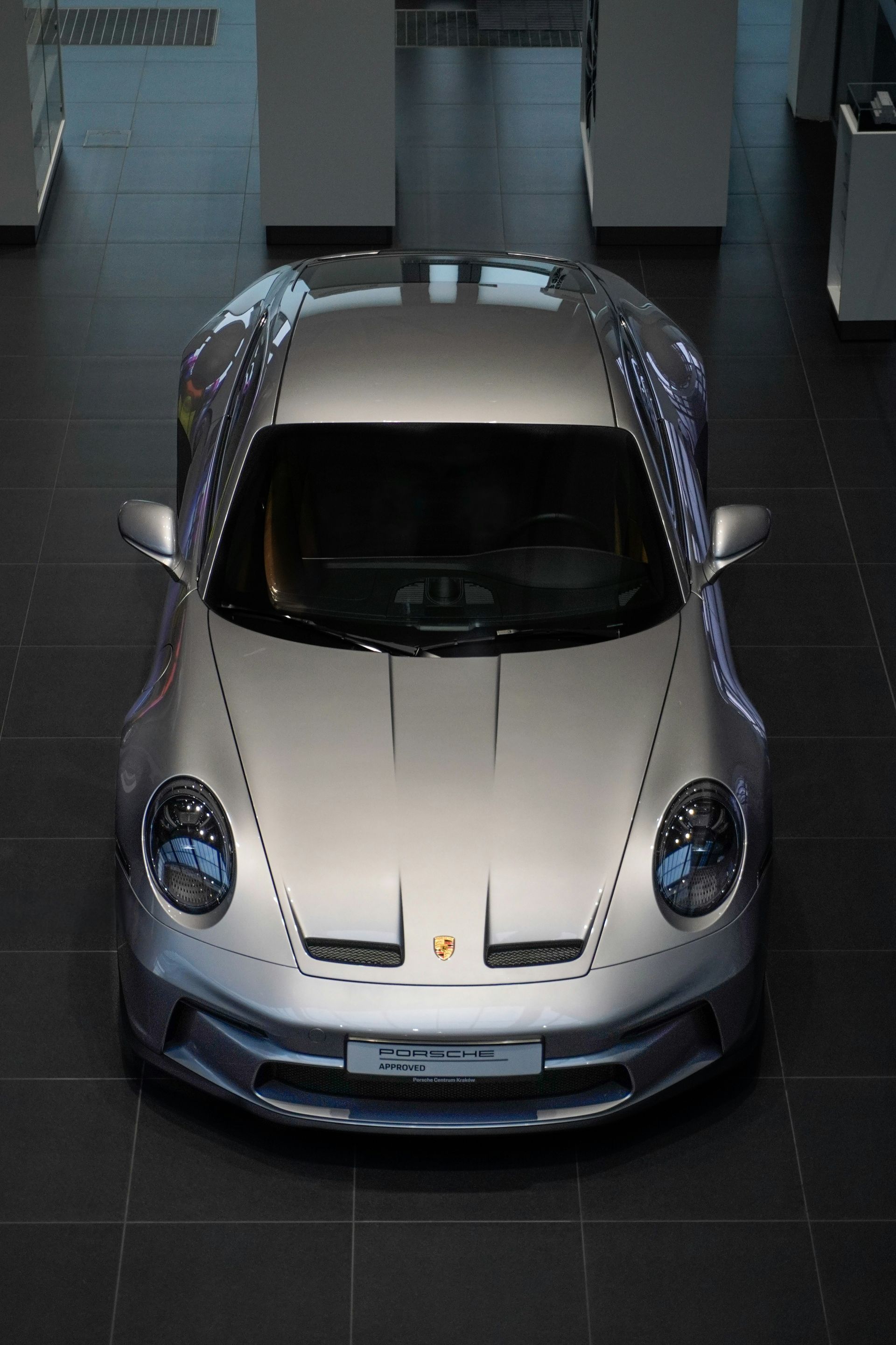 A high-angle view of a silver Porsche 911 parked inside a modern, minimalist showroom with dark gray tiled flooring.