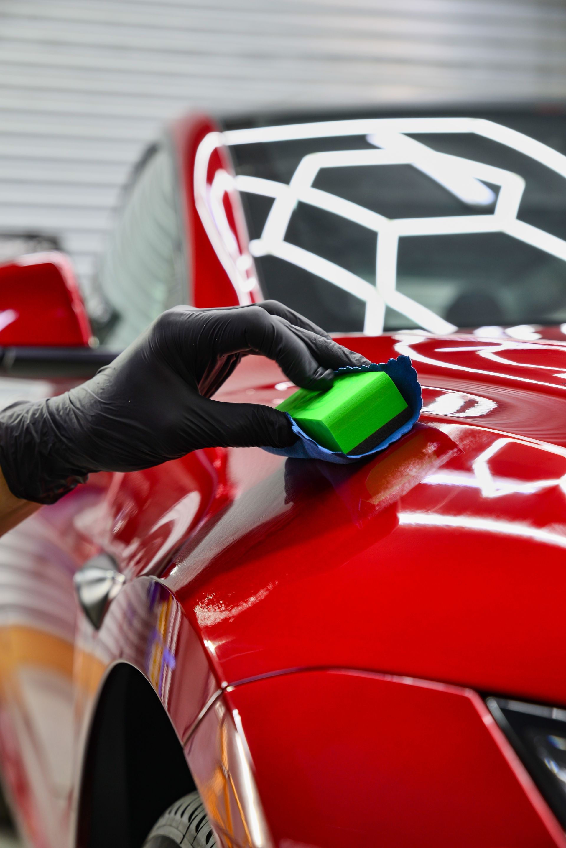 A person in a black glove uses a foam applicator to apply a ceramic coating to the red hood of a car.