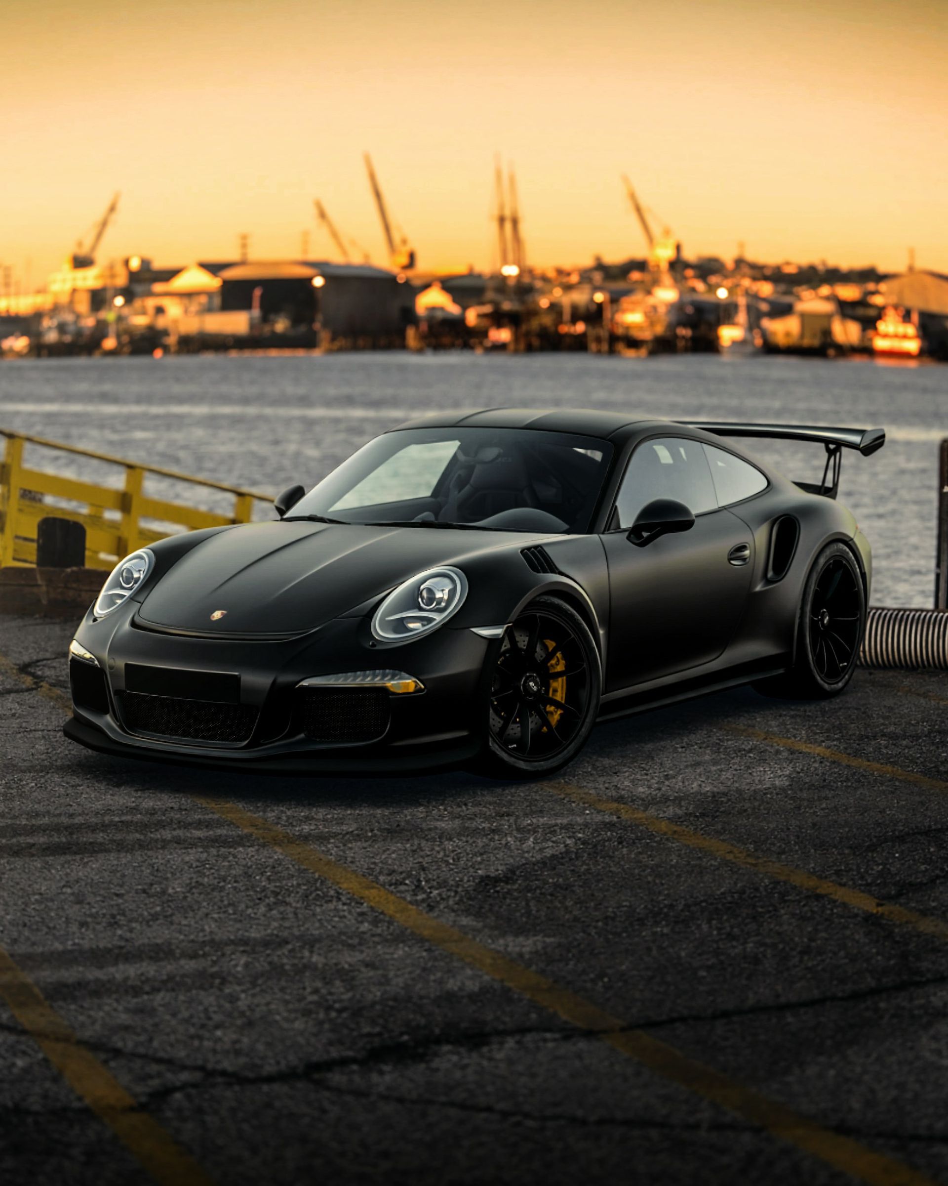 A matte black Porsche sports car parked on asphalt by a harbor at sunset, with cranes and ships in the background.