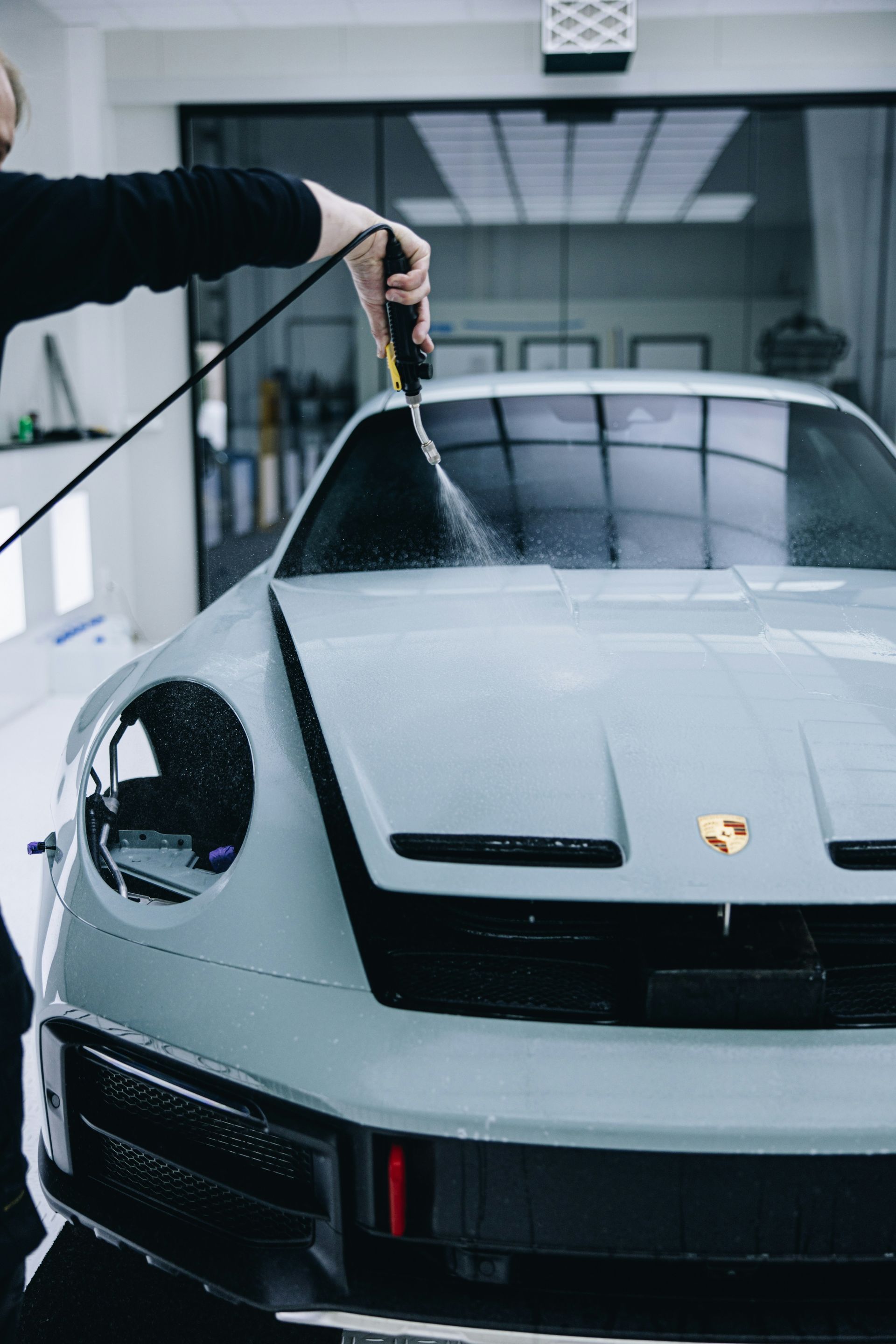 A close-up of a person using a pressure washer to clean a light grey Porsche in a bright, modern auto shop.