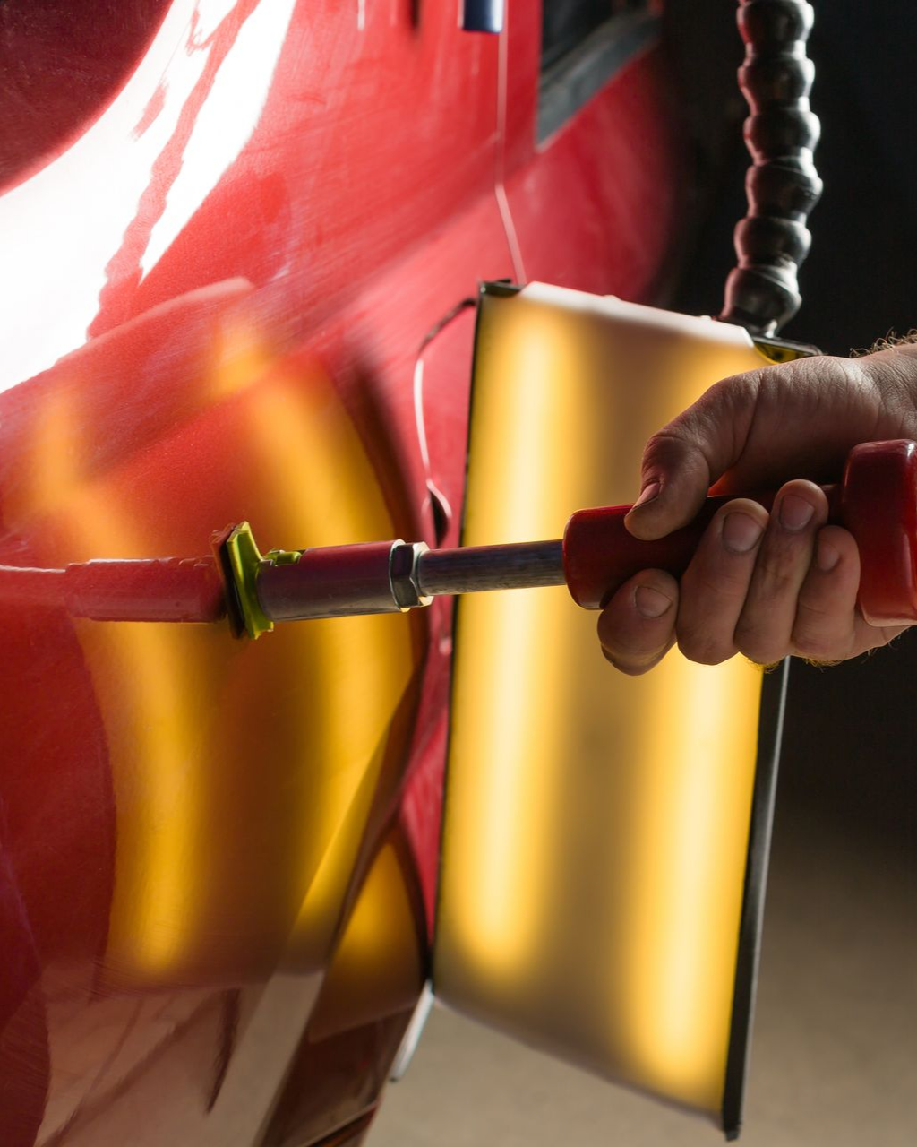 A technician uses a bright work light to inspect small dents on the hood of a black car in a repair shop.