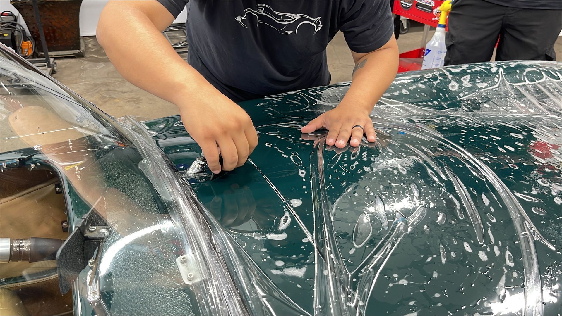 A person installs a clear paint protection film over the hood of a dark green car in an automotive shop.