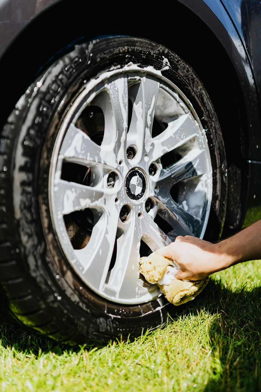 A person uses a yellow sponge to scrub suds off a silver car wheel over a grassy lawn.