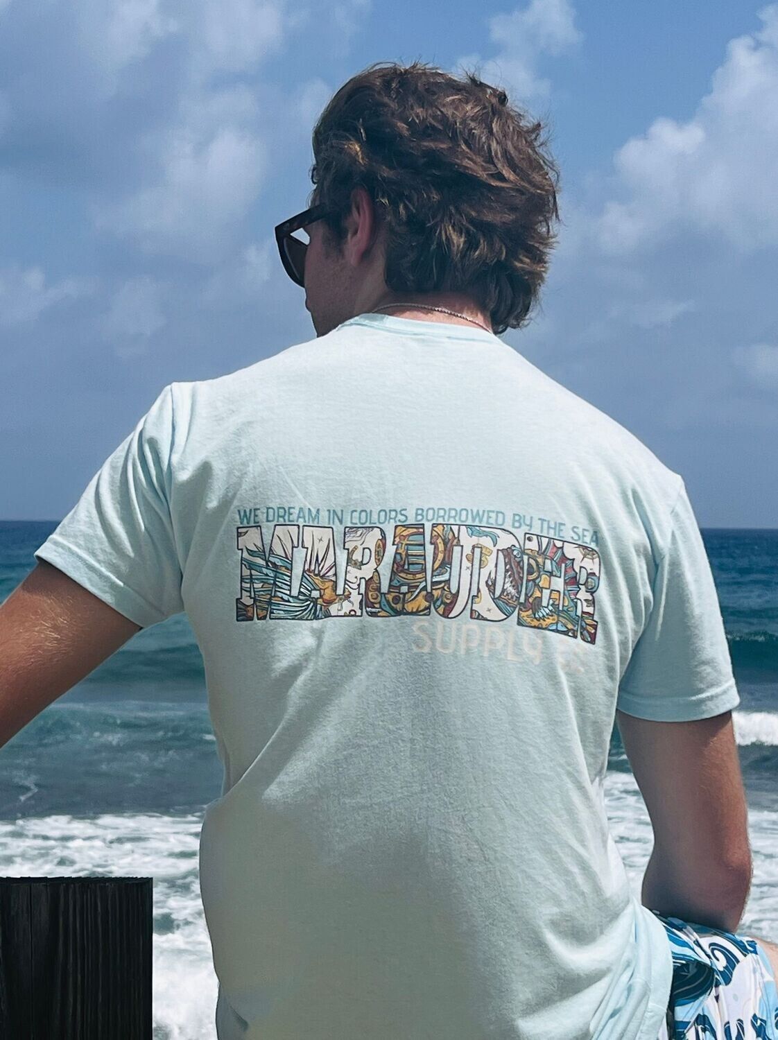 A man wearing a light blue t-shirt is sitting on the beach