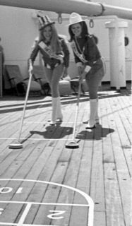 Two people playing deck shuffleboard, leaning on cues on an outdoor ship deck.