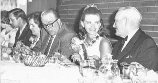 Formal dinner guests seated at a long table, including a smiling woman and an elderly man in suits.