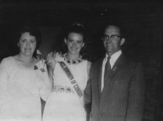 Three people posing for a formal black-and-white portrait, with the center person wearing a sash and corsage.