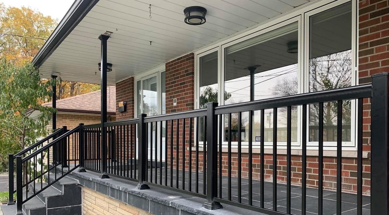 A brick house exterior features a covered porch with black metal railings and steps, set against trees in the background.