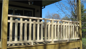 Light-colored wooden porch railing with alternating vertical slats against a dark house and blue sky.