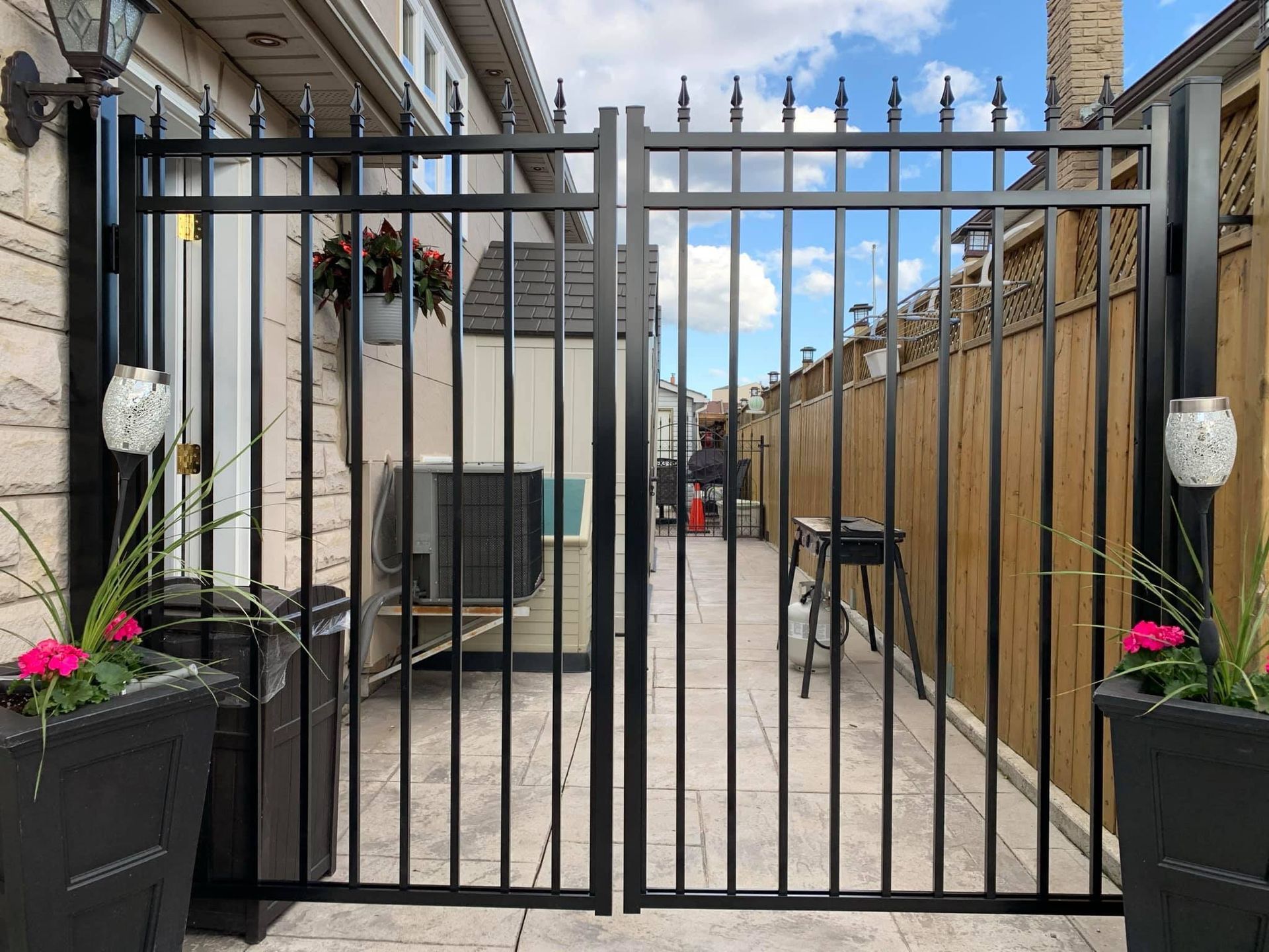 A black metal double gate stands between a stone house wall and a wooden fence, flanked by two planters with pink flowers.