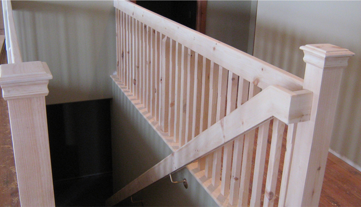 Unfinished light-colored wooden railing and stairwell with vertical balusters and two square posts.