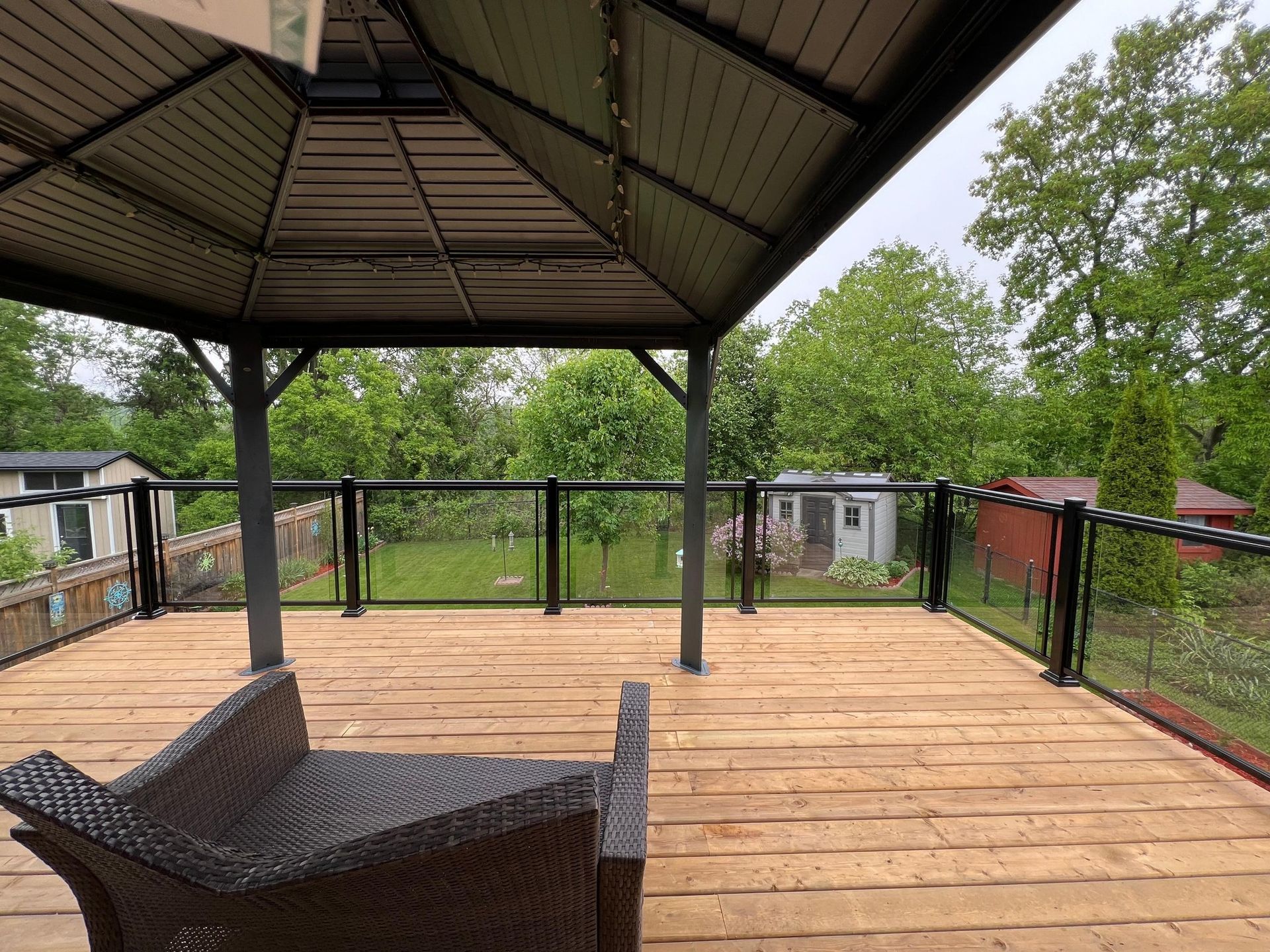 A wooden deck featuring a dark gazebo over a wicker lounge chair, overlooking a grassy yard with small sheds and trees.