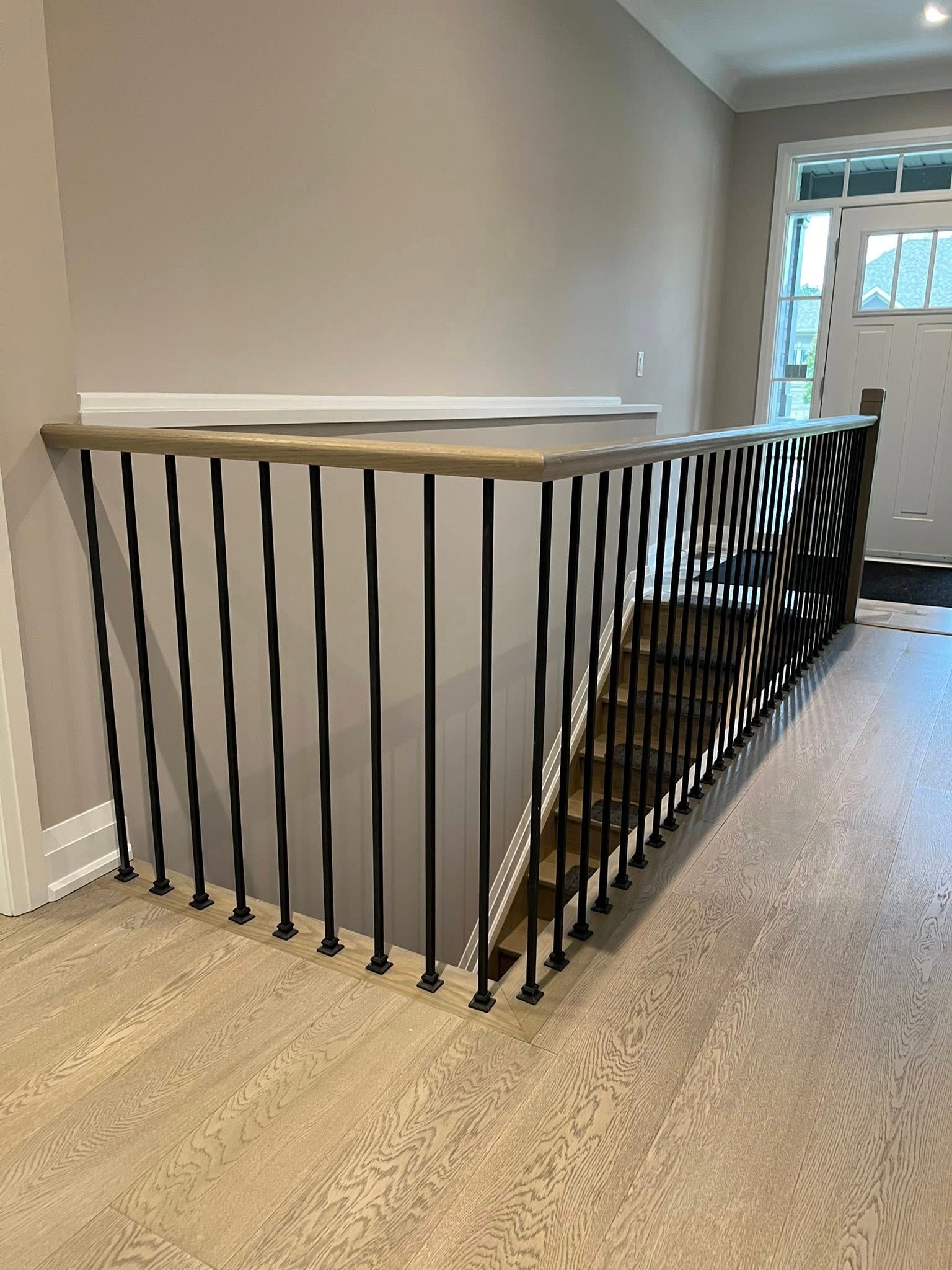 A modern interior view of black, wavy metal stair balusters with a light wood handrail above light oak hardwood flooring.