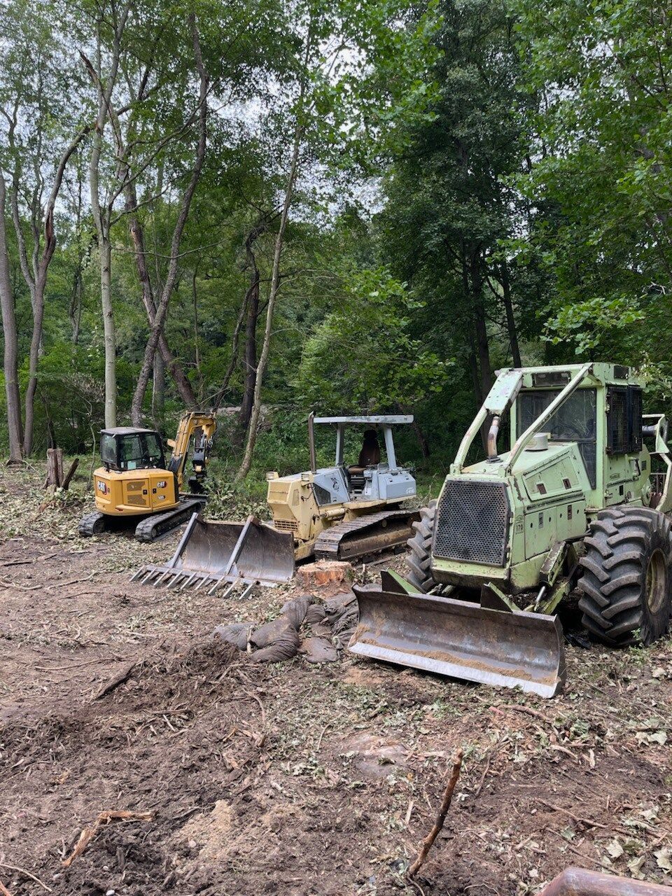 Three pieces of heavy machinery clearing a forest area: excavator, bulldozer, and forestry machine.