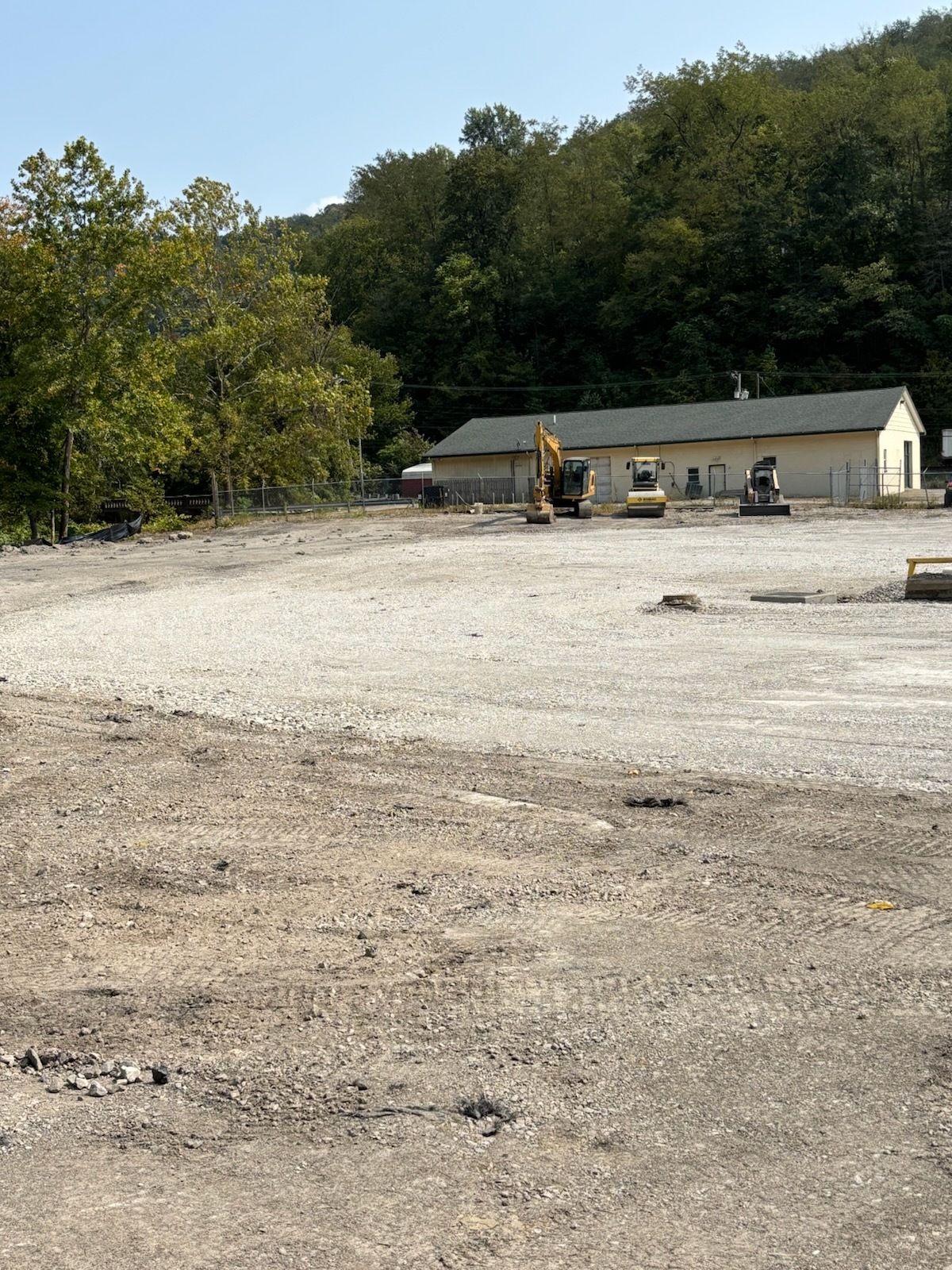 Gravel lot with heavy equipment in front of a single-story building, forested hillside in the background.