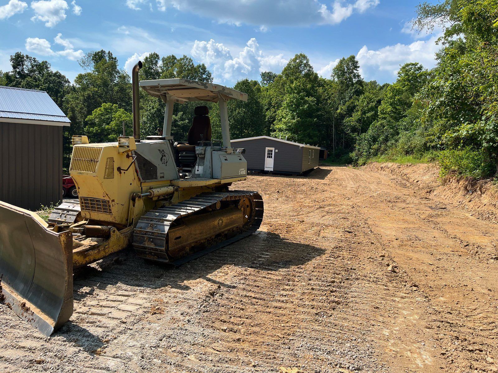 Yellow bulldozer parked on a dirt road near trees and a shed on a sunny day.