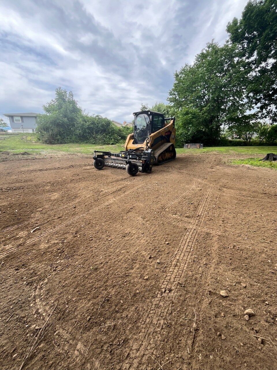Skid steer with land leveler on a dirt lot. Cloudy sky, trees, and a building in the background.