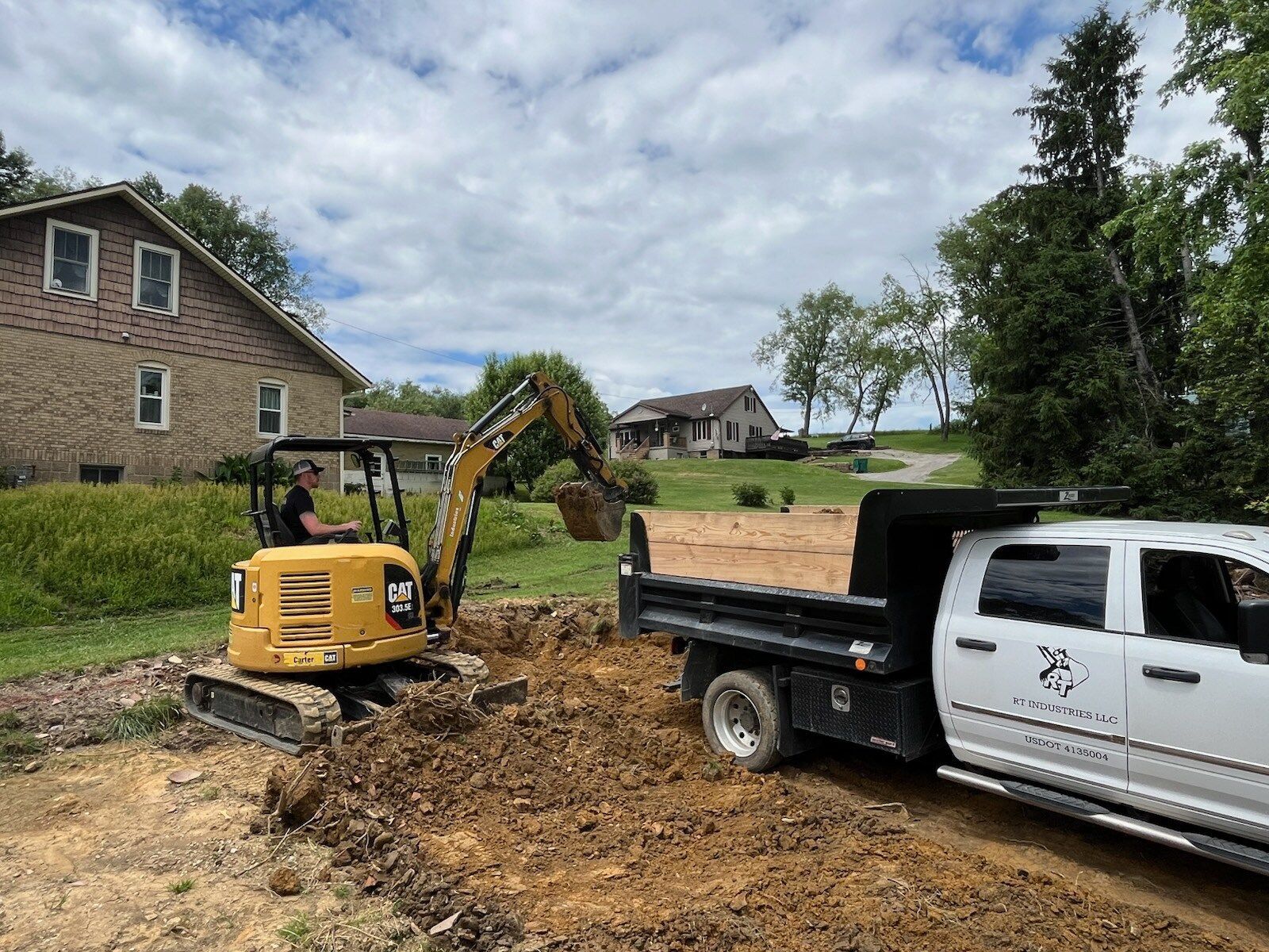 A yellow excavator loads dirt into the bed of a white pickup truck near a house on a sunny day.