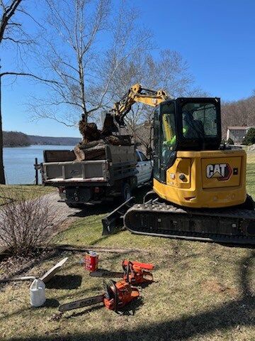 Yellow excavator loading logs into a dump truck near a lake, with two chainsaws on the ground.