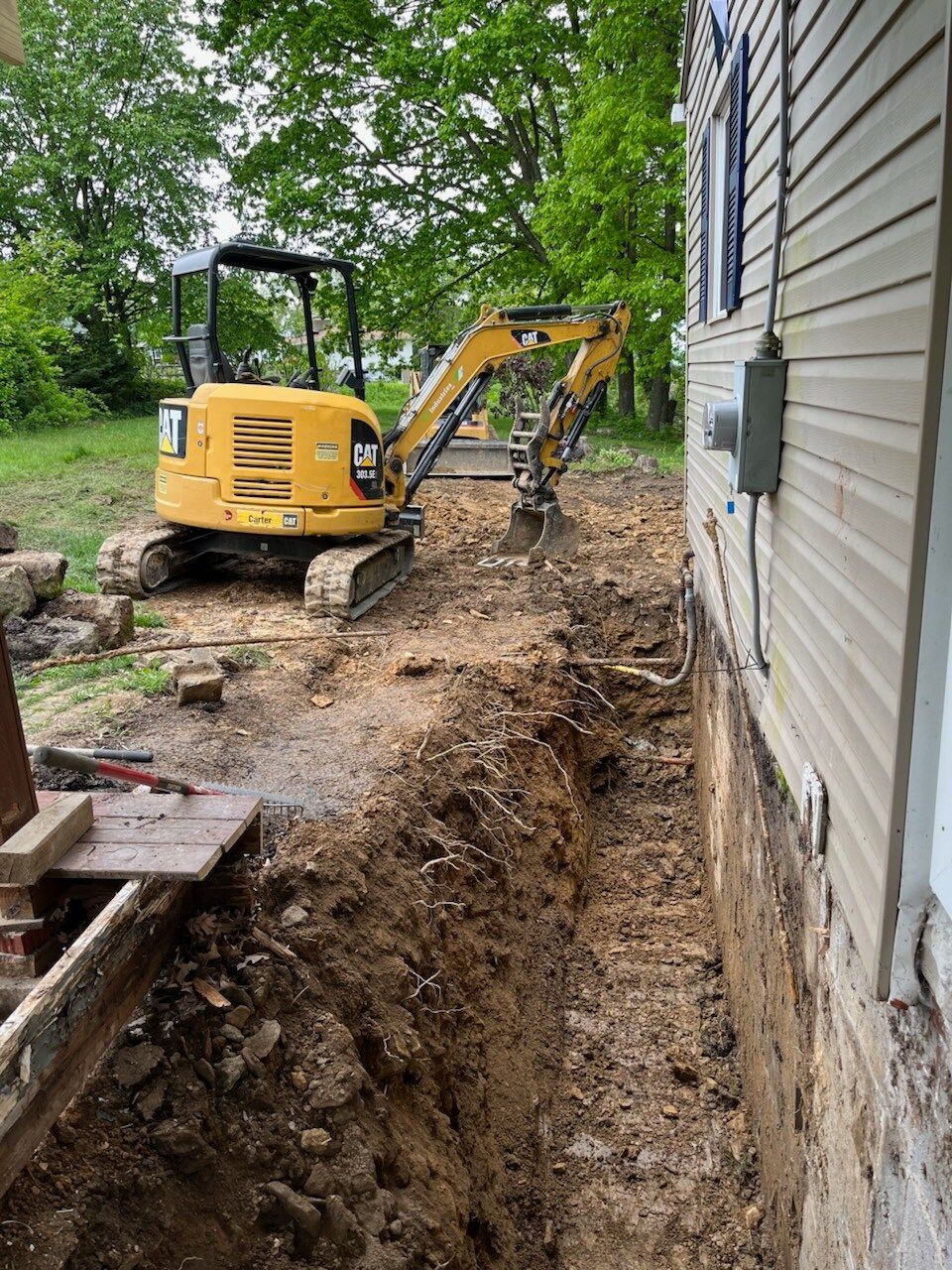 A yellow excavator digging a trench next to a house. Earth is piled up.