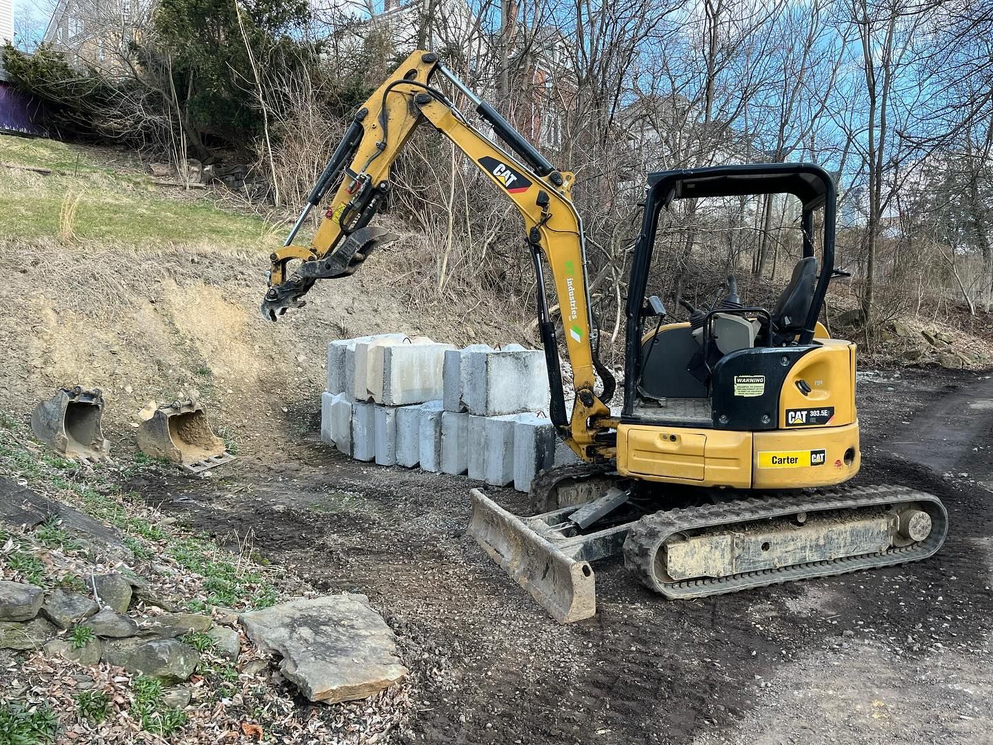 A small yellow excavator is sitting in a dirt field next to a pile of concrete blocks.