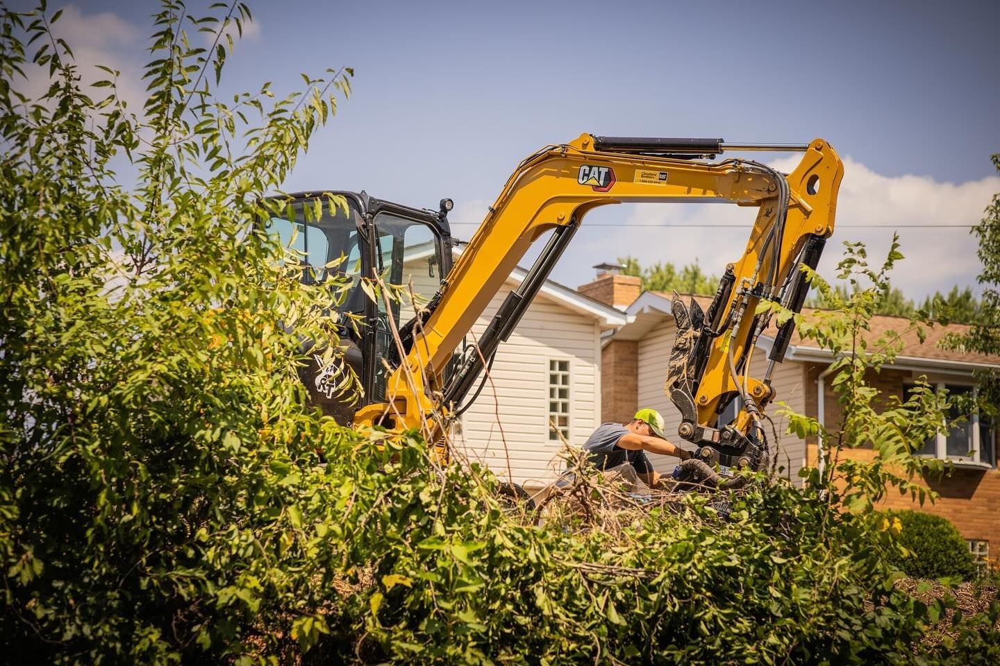 A yellow excavator is cutting a hedge in front of a house.