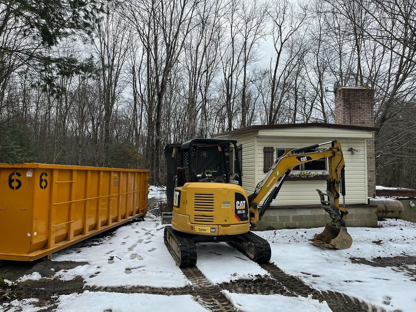 A yellow excavator is parked in front of a house in the snow.