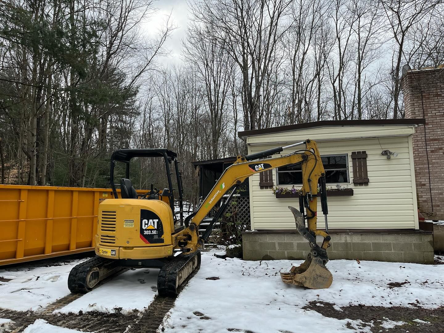 A yellow excavator is parked in front of a house in the snow.