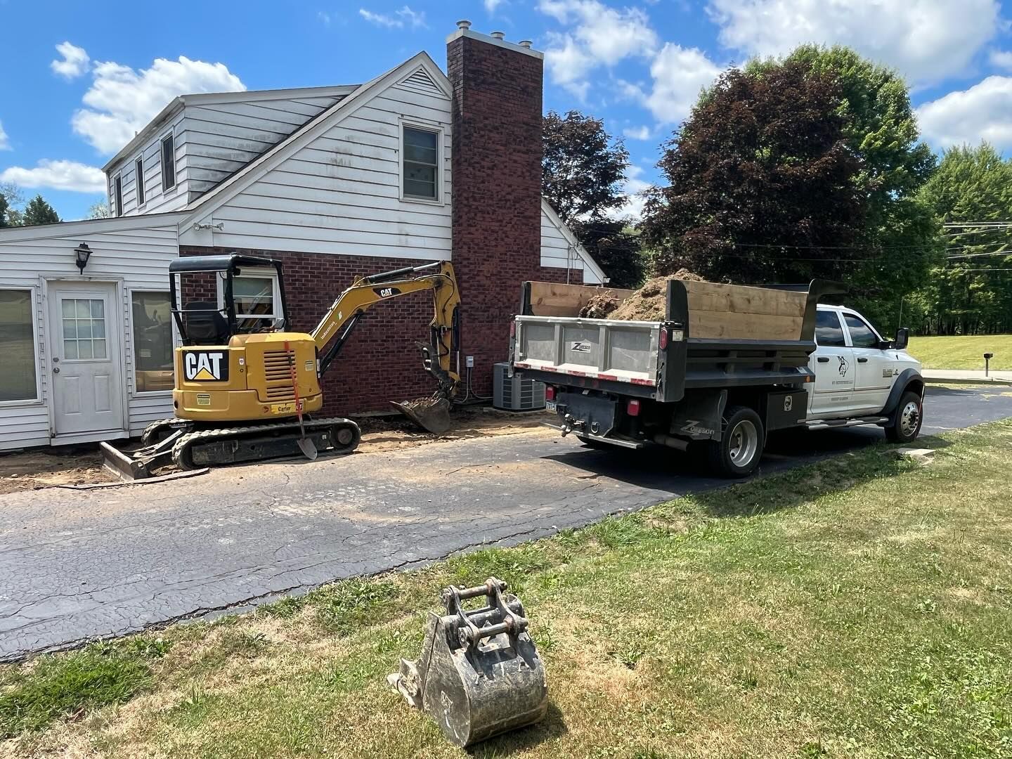 A small excavator is parked in front of a house next to a dump truck.
