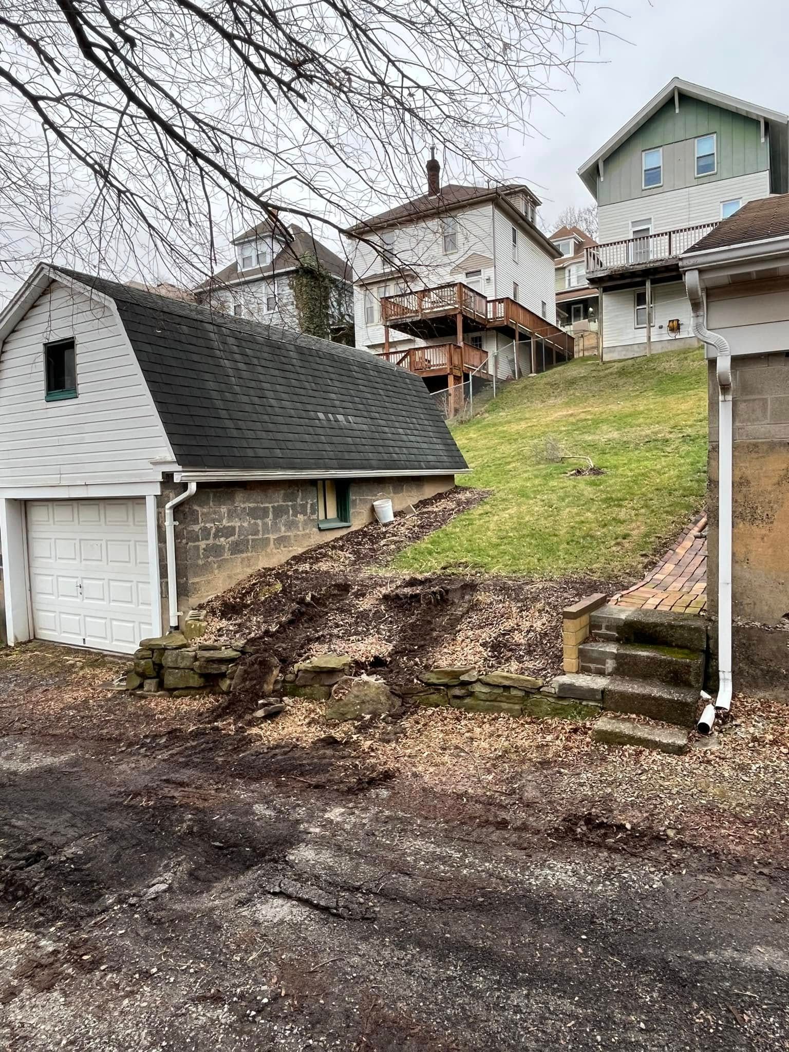 A white house with a black roof is sitting on top of a hill next to a garage.