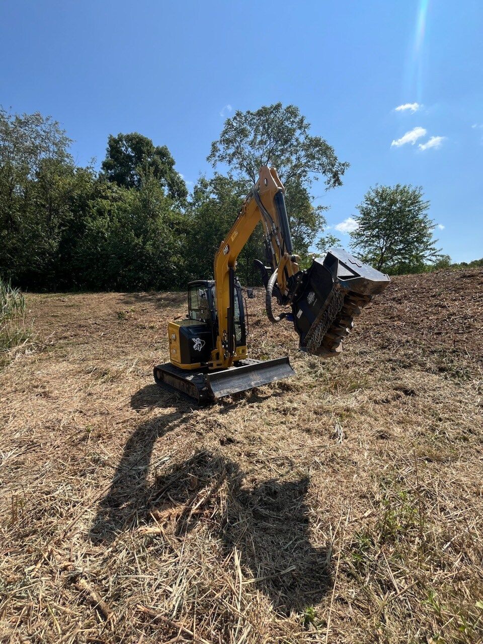 Yellow excavator with a mulching head clearing brush in a field on a sunny day.