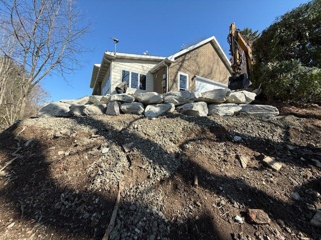A house on a hillside with large rocks forming a retaining wall, an excavator is in the background.