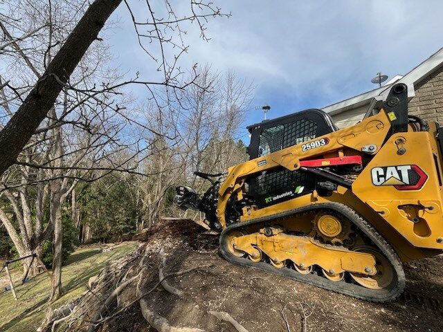 Yellow Caterpillar track loader on a hillside, near a house and trees, clearing dirt.