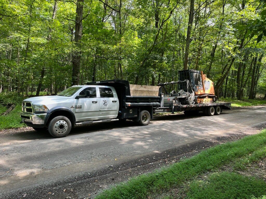 White work truck hauling heavy machinery on a gravel road in a wooded area.