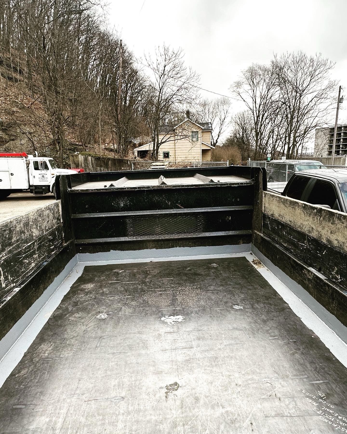 Empty truck bed, grey-lined, against a backdrop of bare trees and buildings on a cloudy day.