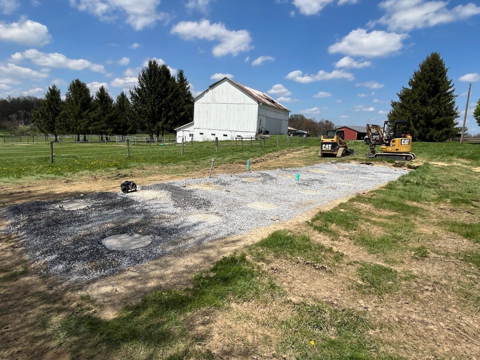 Construction site with gravel base, two mini excavators, white barn, and blue sky.