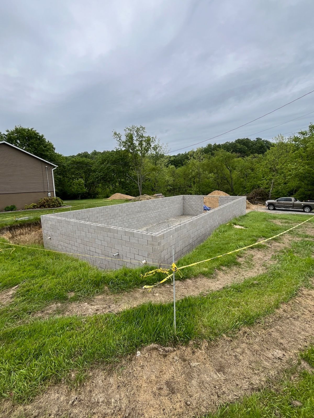 Concrete foundation of a building under construction, yellow caution tape, grass, trees, overcast sky.