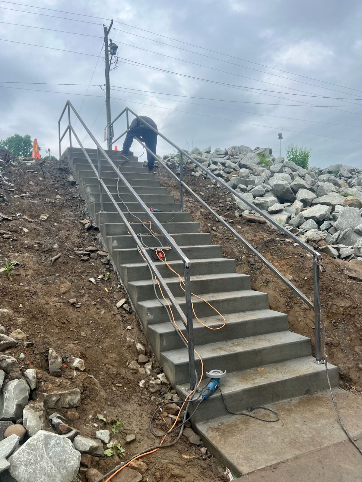 Person working on metal railings of concrete stairs on a slope. Gray sky and rocky ground.
