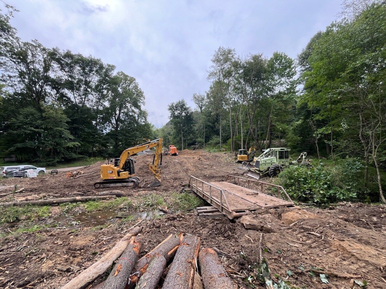 Construction site in a forest with heavy machinery, cut logs, and a makeshift bridge.