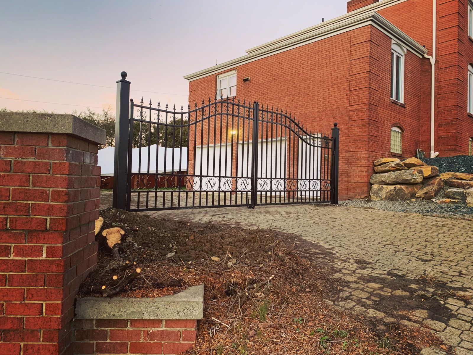 Black iron gates on a brick driveway lead to a red brick building with white doors.