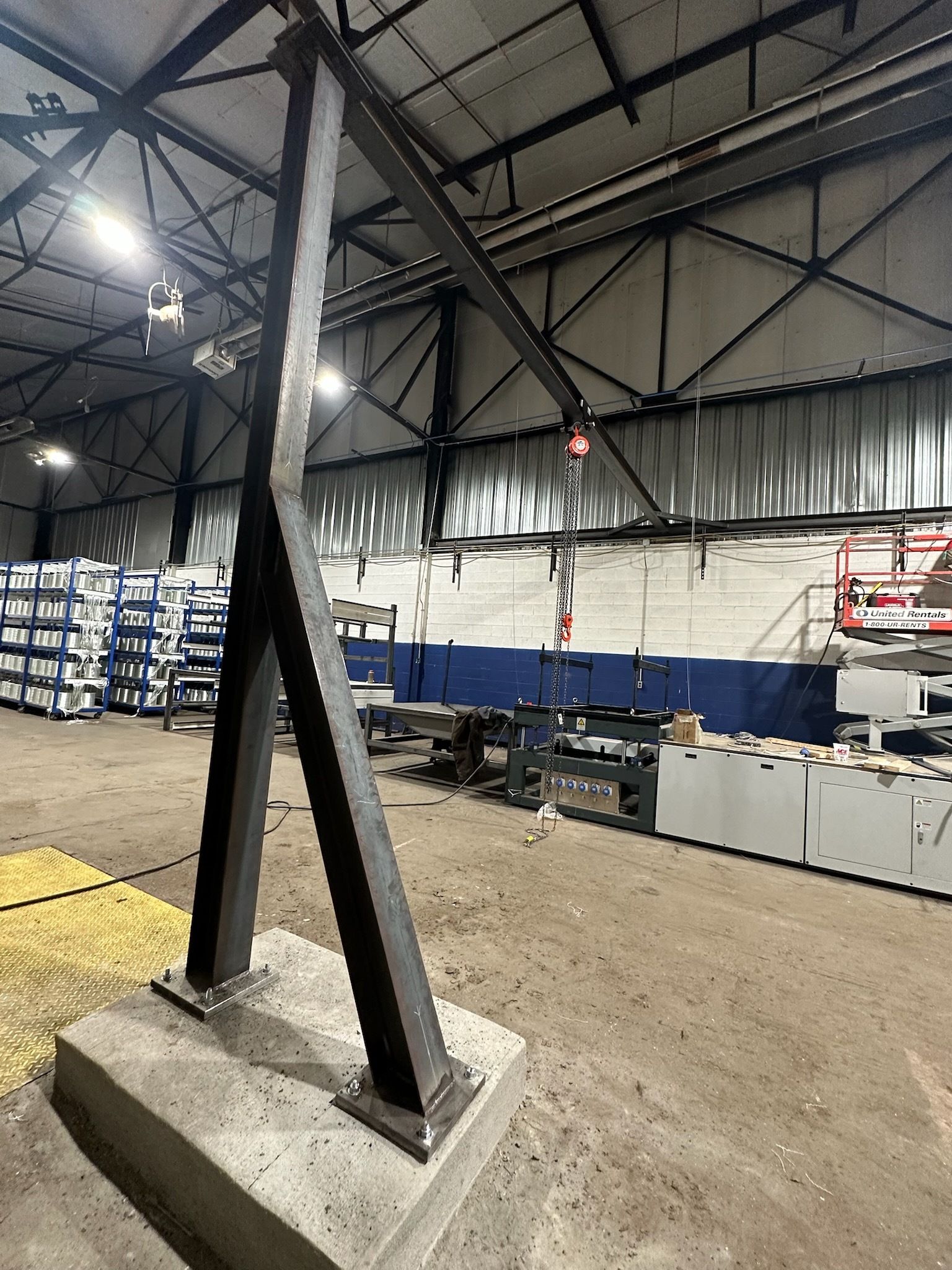 A-frame crane in a warehouse setting. Black metal construction, red hook, concrete base.