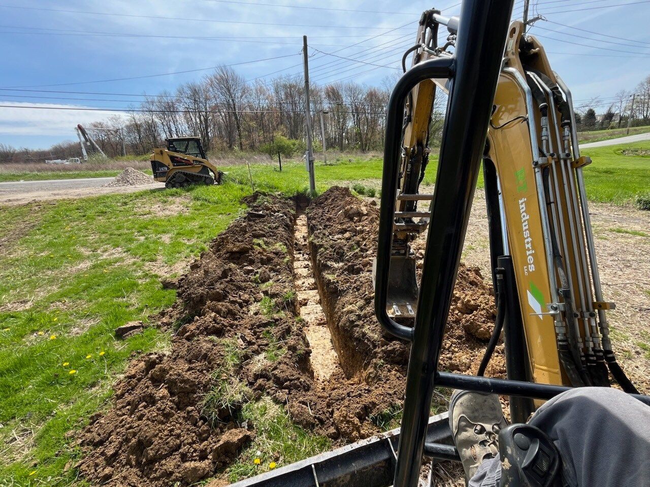 A trench being dug by a mini excavator in a grassy area with a small tractor in the background.