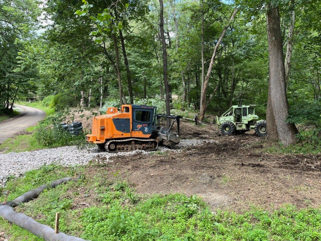 Orange and green construction vehicles in a wooded area, working on a gravel path near trees.